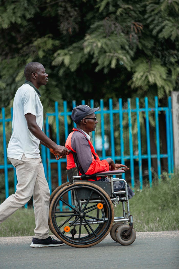A man pushes another man in a wheelchair along a city street by a blue fence.